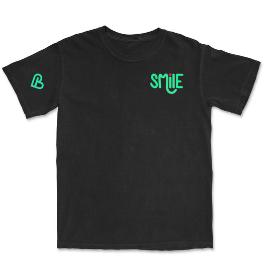 A black t-shirt laid flat, featuring a vibrant green and pink "SMILE" logo on the left chest area. The design has a playful, slightly bubbly font with the "I" dotted by a small circle.  A heart shaped PSTVE Brand emblem on the right sleeve.